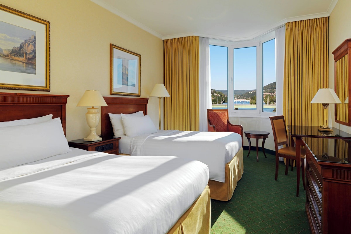 A hotel room with two beds, a desk, a chair, a small table, and a view of the outdoors.