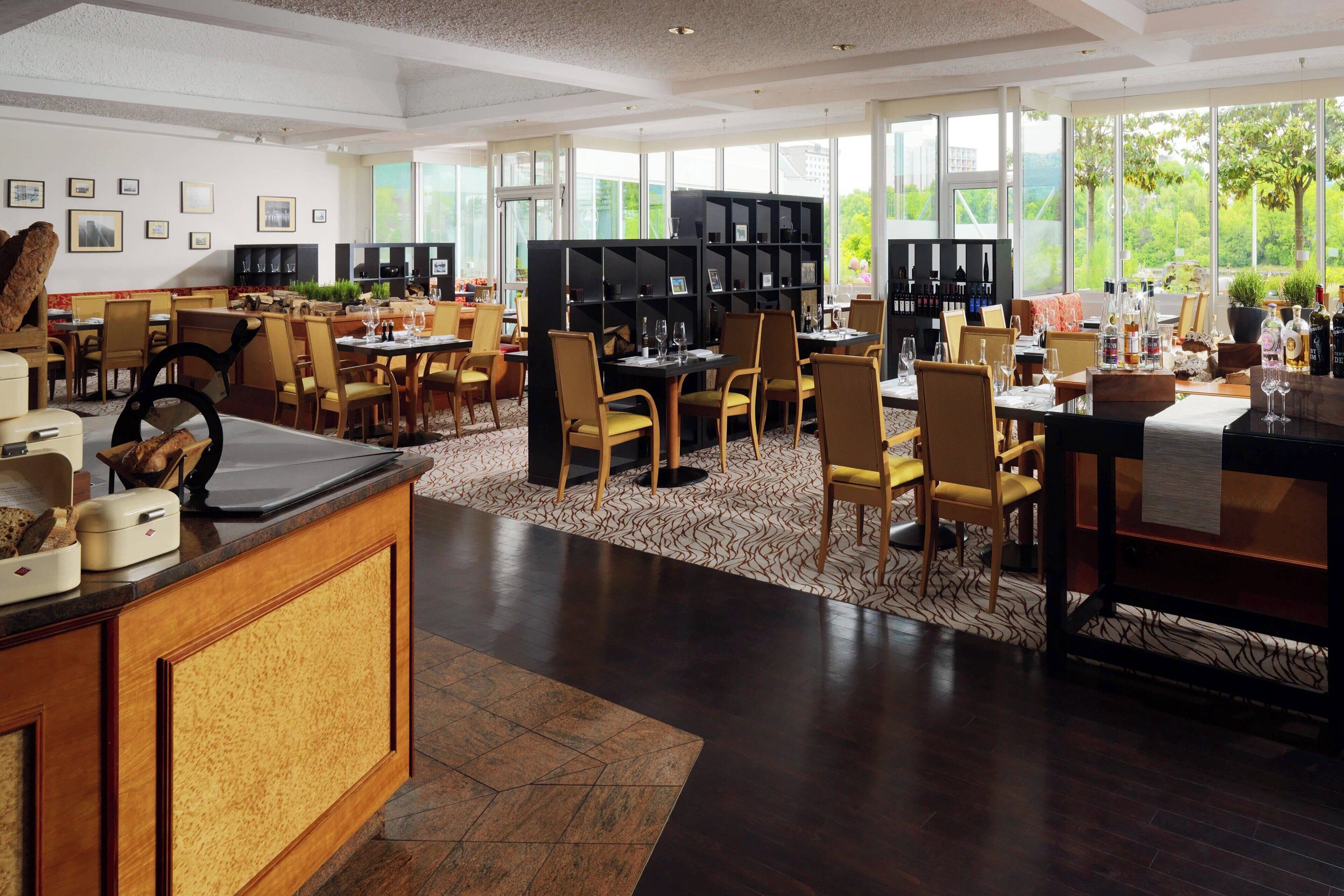 A spacious hotel dining area with multiple tables and chairs, a central black bookshelf, and large windows offering a view of the outdoors.