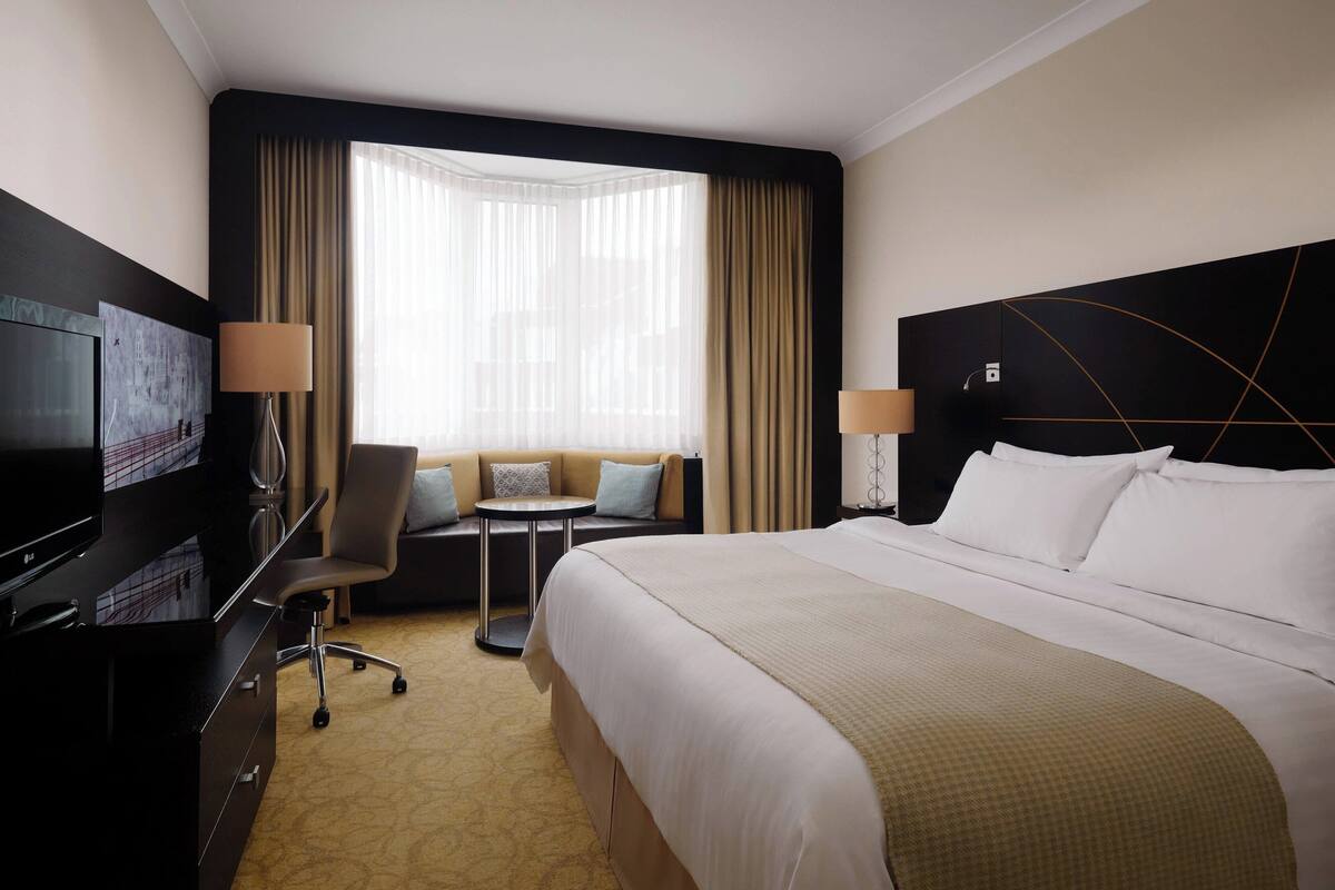A hotel room with a large bed, a desk with a TV, a small table with chairs, and a window with curtains.