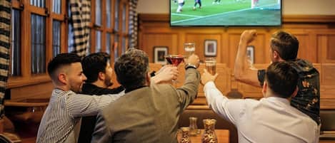 A group of people watching a soccer match on a television screen in a room with wooden paneled walls and a bar area.