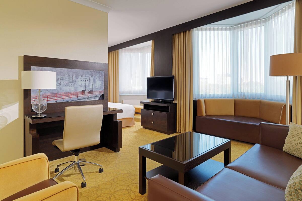 A hotel room with a desk, a television, a sofa, and a large window with curtains.