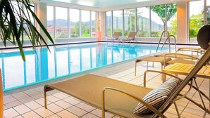 Una piscina interior con agua azul cristalina, rodeada de grandes ventanales que ofrecen vistas a zonas verdes y edificios. Hay sillones de descanso con cojines a lo largo de la piscina.