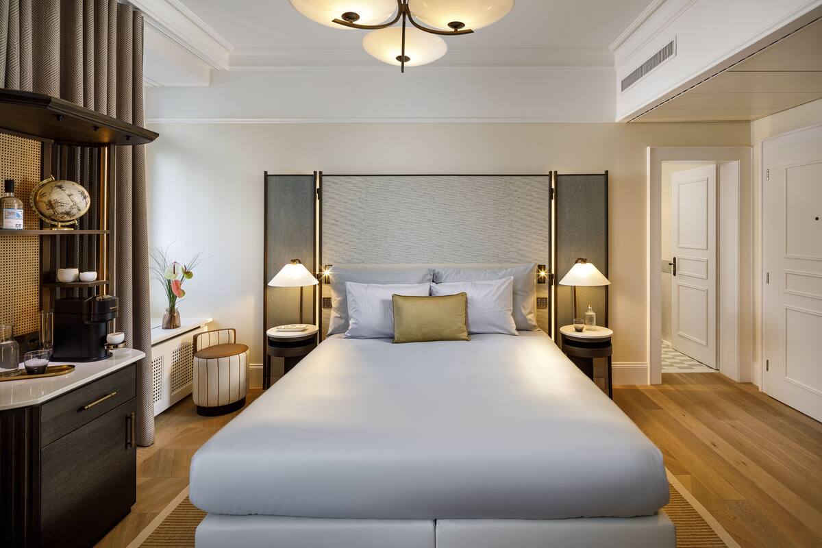 A spacious hotel room with a large bed, two bedside tables with lamps, a small side table, a globe, and a minibar.