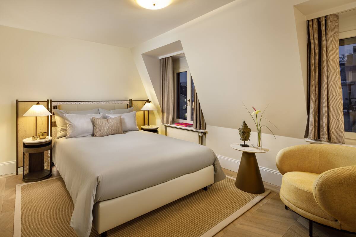 A modern hotel room with a large bed, two bedside tables with lamps, a small side table with a vase, a chair, and a window with curtains.
