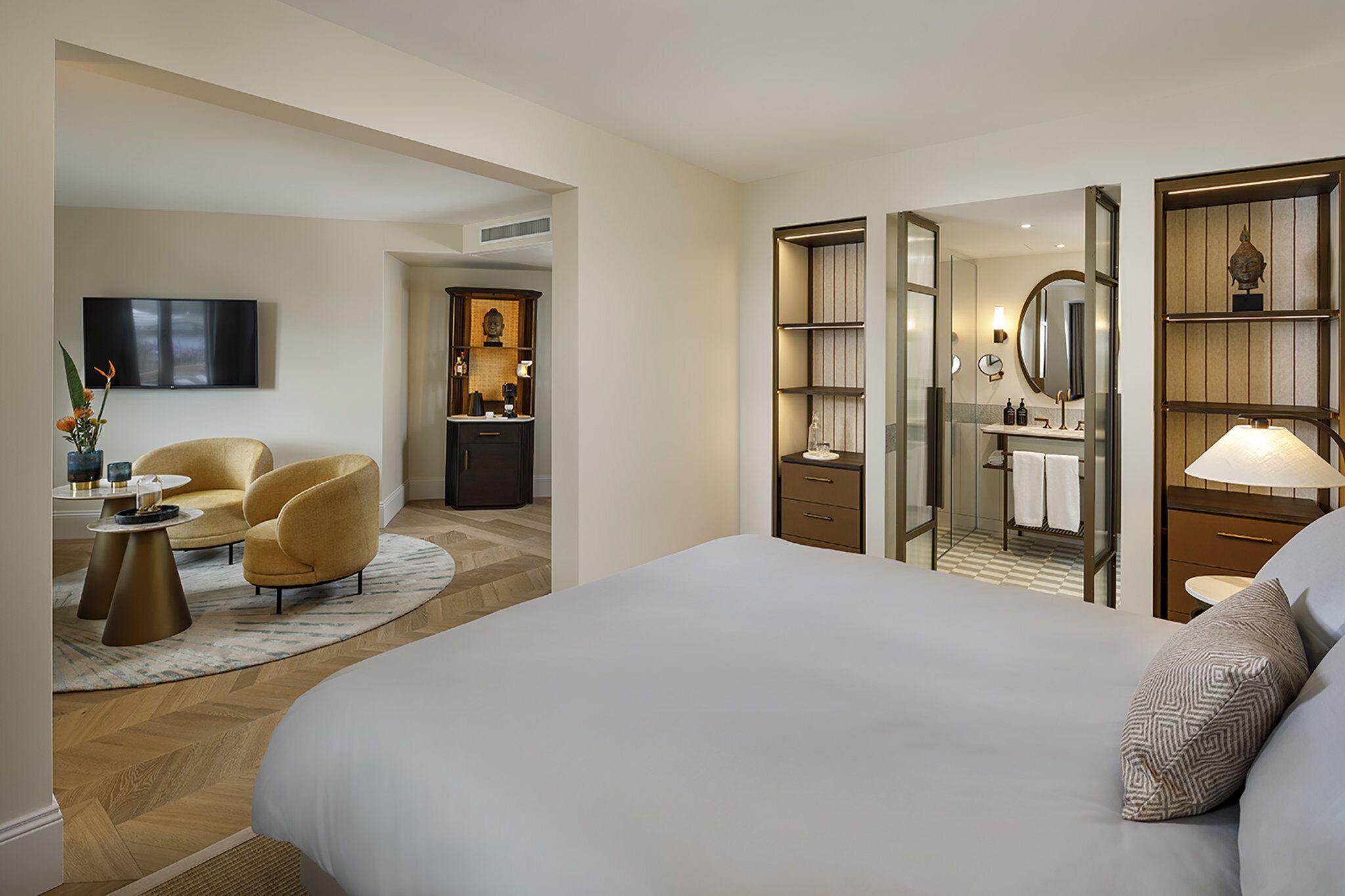 A hotel room with a large bed, a seating area with two armchairs, a TV, a desk with a lamp, and a bathroom visible through a glass door.