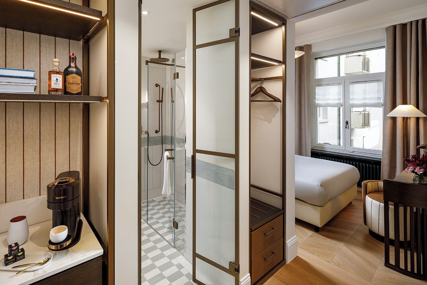 A modern hotel room with a walk-in closet, a separate bathroom with a glass shower, a bed, a dining table with a lamp, and a view of the outside through large windows.