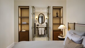 A hotel room with a large bed, a bedside table with a lamp, a wardrobe with shelves, a mirror, and a bathroom visible through an open door. - Small Luxury Hotel Ambassador Zürich (Zürich)