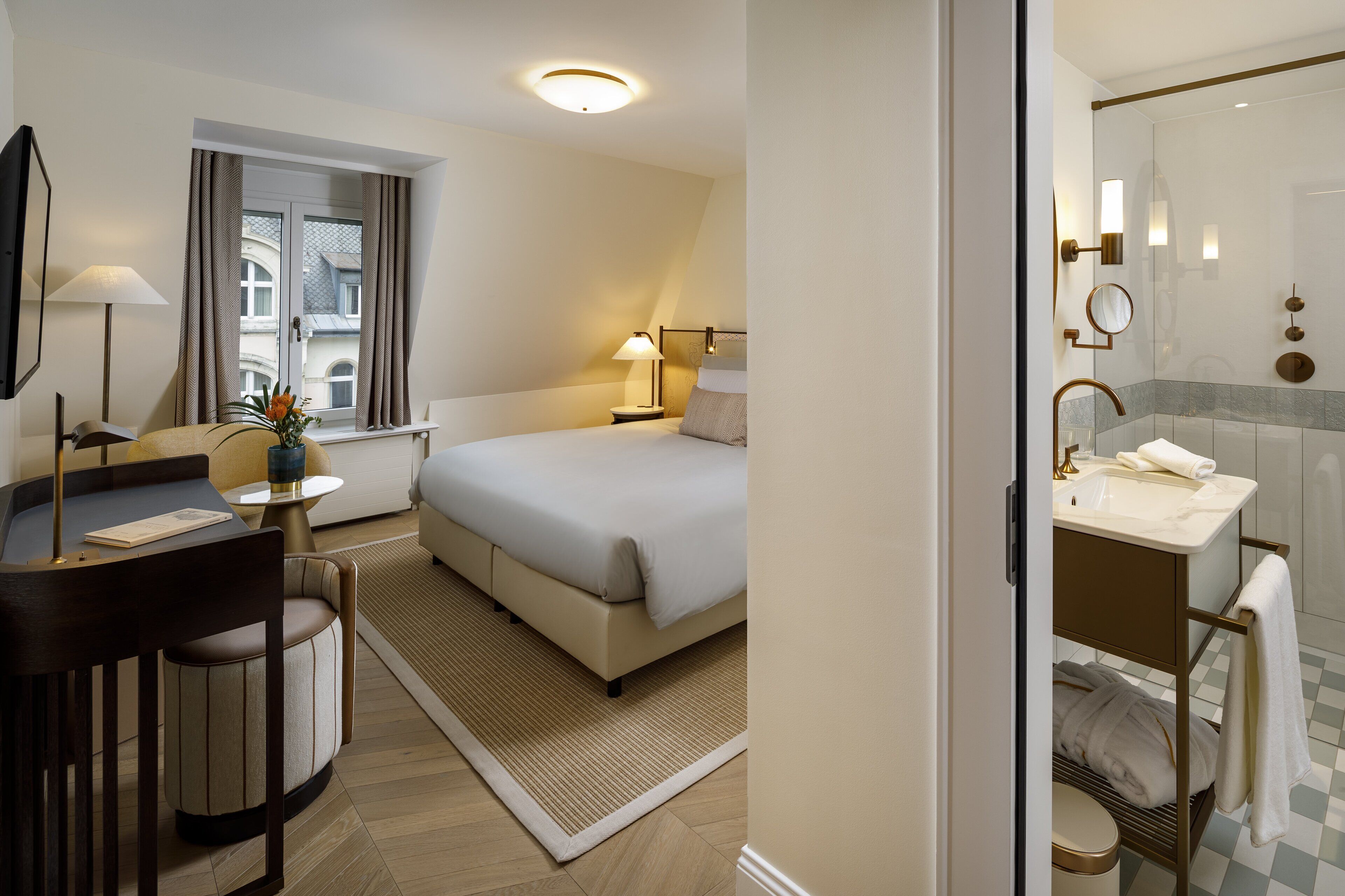 A hotel room with a bed, bedside table, lamp, and a view of the cityscape through a window.