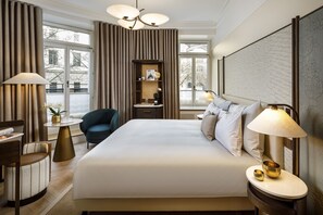 Habitación de hotel con una cama grande, un escritorio con lámpara, una silla y vistas al exterior a través de amplias ventanas.
