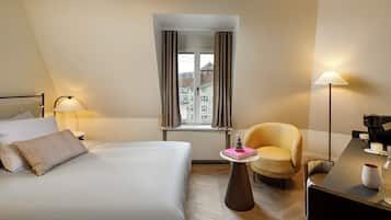 Et hotelværelse med en seng, en stol, et bord med lampe, kaffemaskine og udsigt til bygninger gennem et vindue med gardiner.