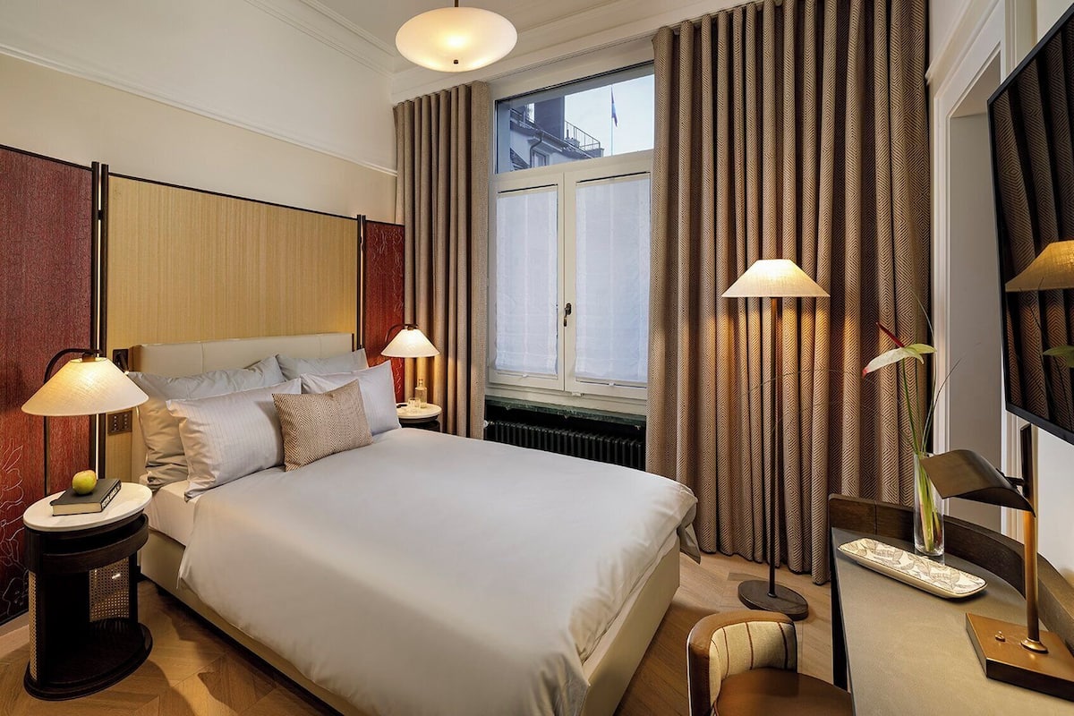 A hotel room with a large bed, two bedside lamps, a nightstand with a book, a small table, and a chair.
