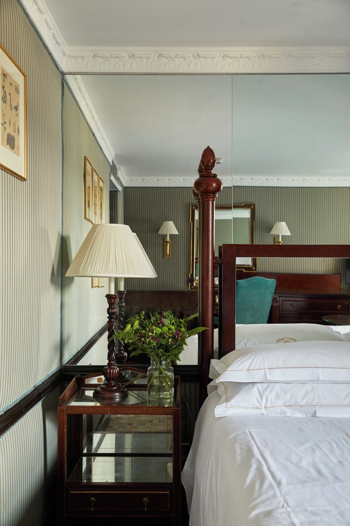 A hotel room with a bed, a nightstand with a lamp, a vase with flowers, and a mirror on the wall.