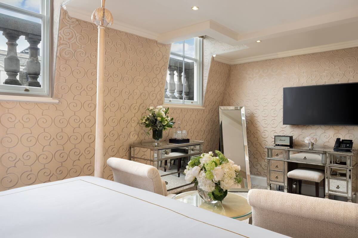 A hotel room with a large bed, a sitting area with a sofa and armchair, a vanity with a mirror, a dresser, and a flat-screen TV mounted on the wall.