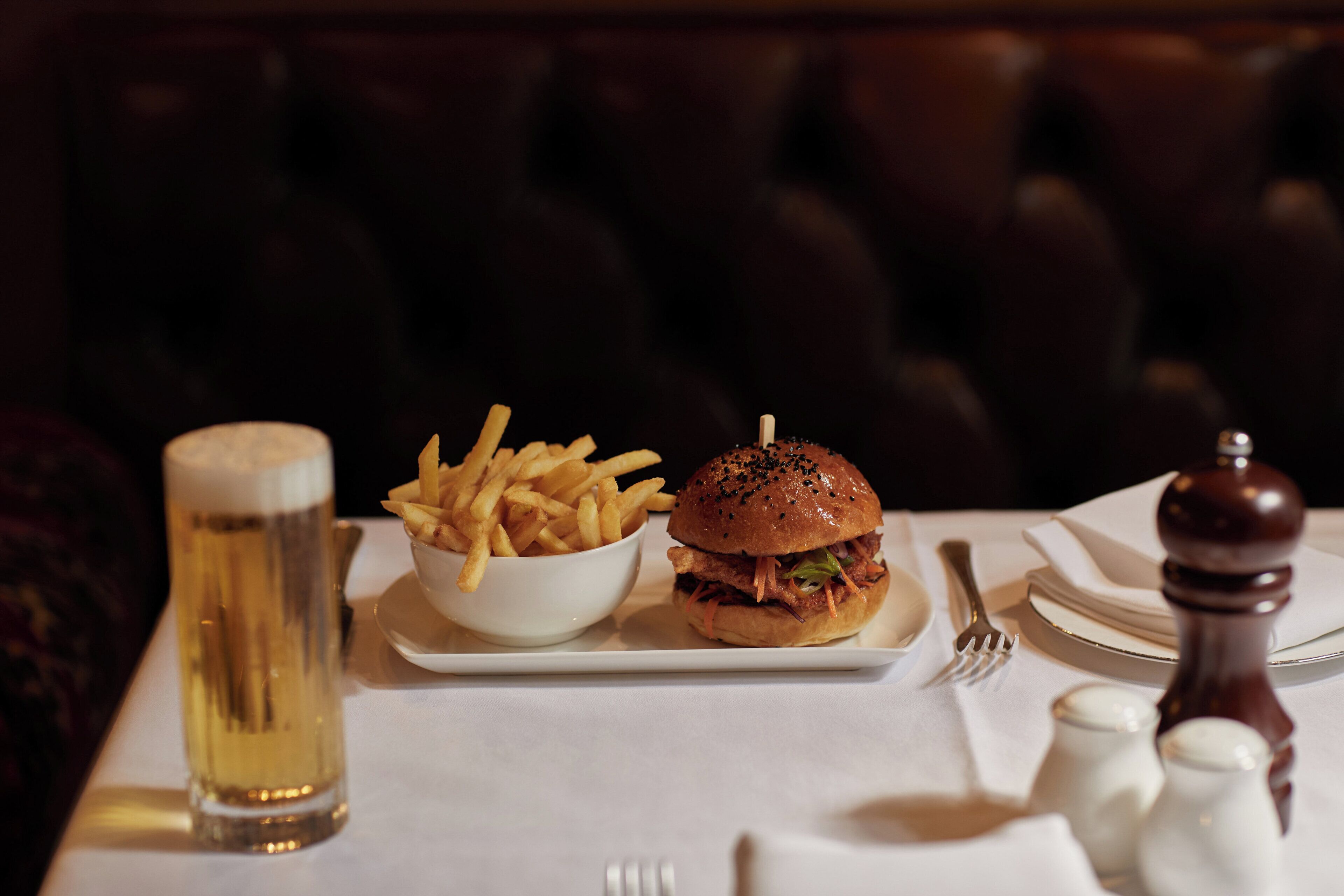 Ein Essen bestehend aus einem Burger und Pommes auf einem weißen Teller, ein Glas Bier sowie Salz- und Pfefferstreuer auf einem Tisch.
