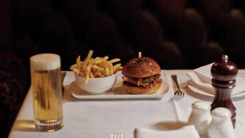 Ein Essen bestehend aus einem Burger und Pommes auf einem weißen Teller, ein Glas Bier sowie Salz- und Pfefferstreuer auf einem Tisch.