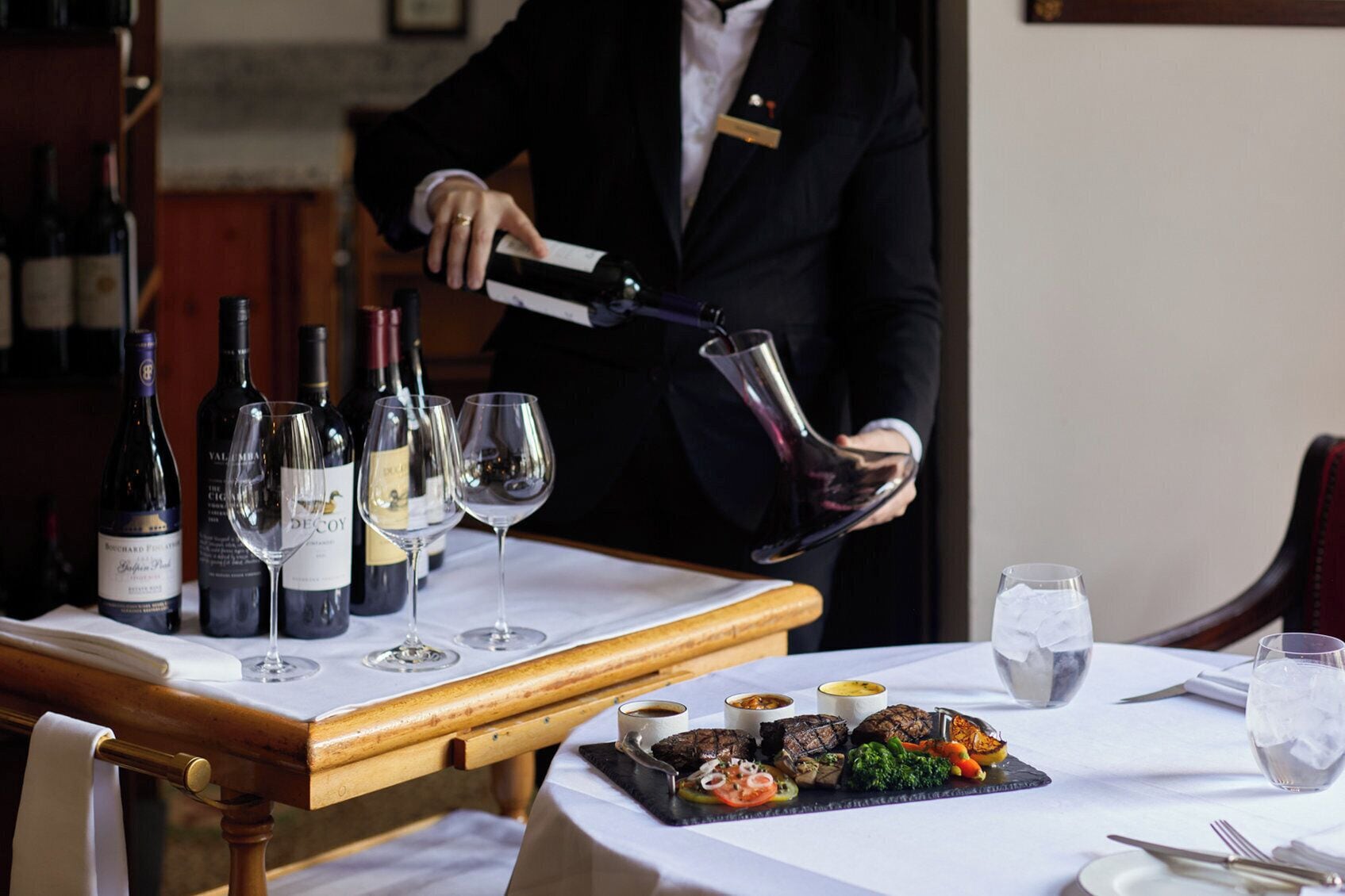 Una persona sirve vino en copas a una mesa, con comida y botellas de vino al fondo.