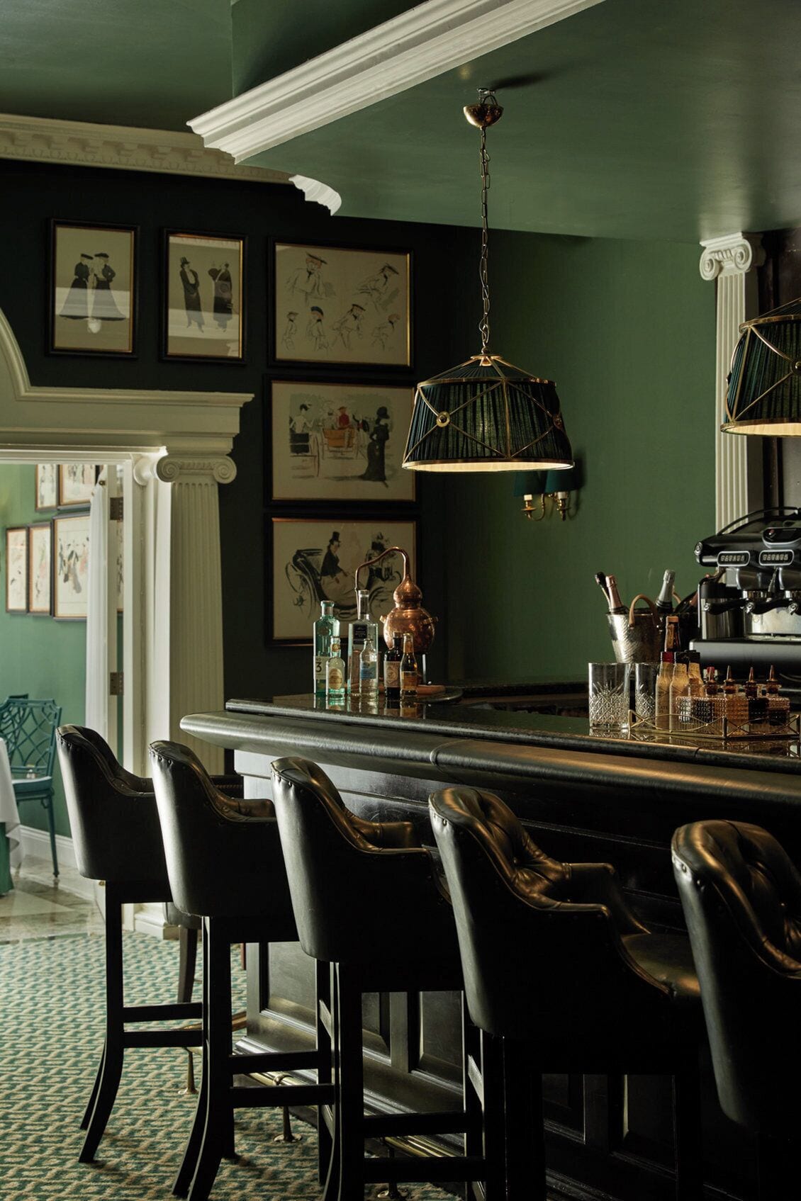 A bar area with a dark green wall, framed artwork, and a hanging light fixture.