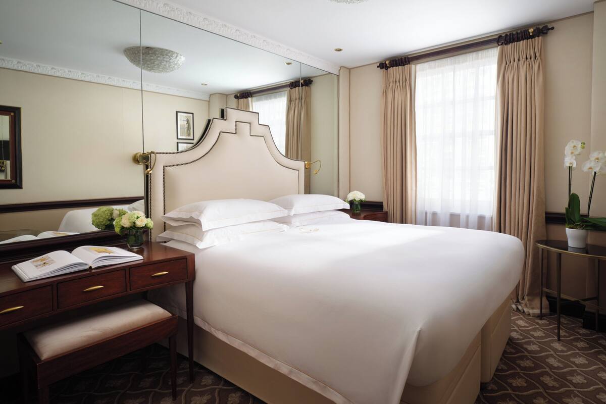 A hotel room with a large bed, a desk with a chair, a bedside table with a vase of flowers, and a mirror on the wall.