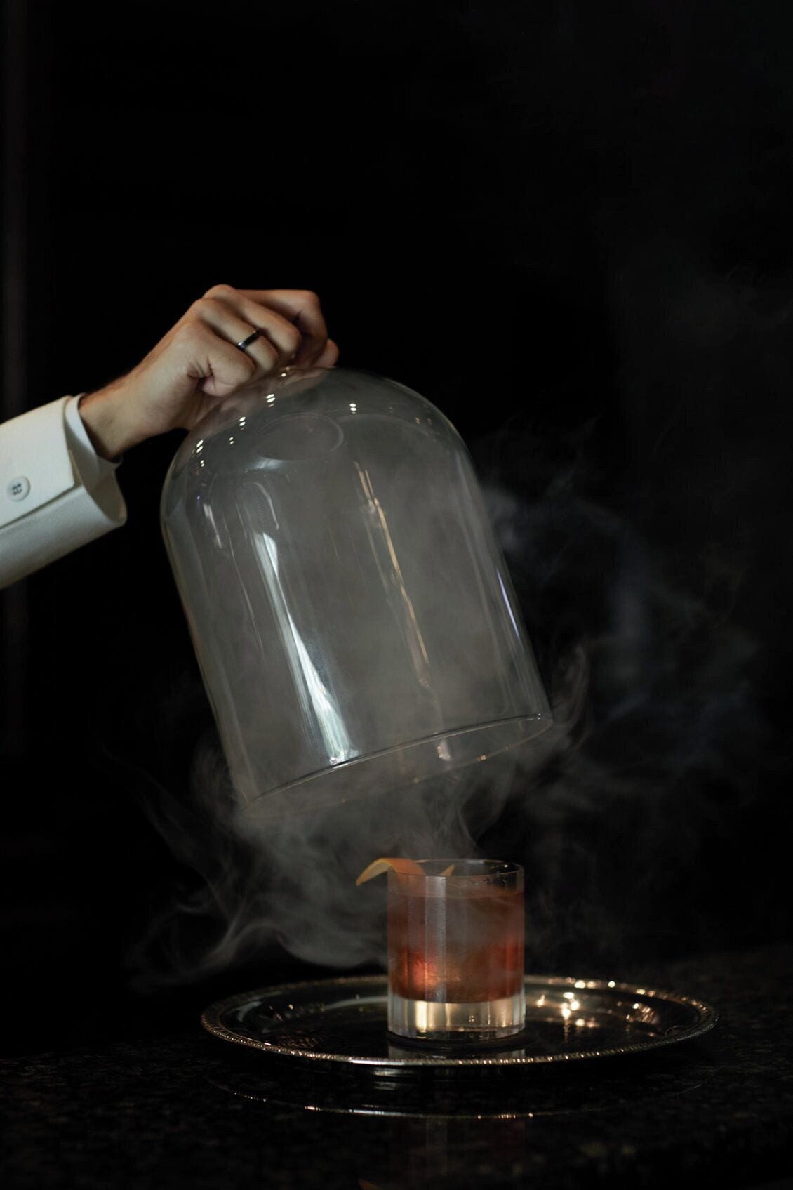 A hand lifting a transparent dome over a glass with a drink, steam rising from the glass.