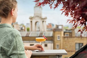 Eine Person hält auf dem Balkon einen Cocktail, im Hintergrund ist ein historisches Gebäude zu sehen.