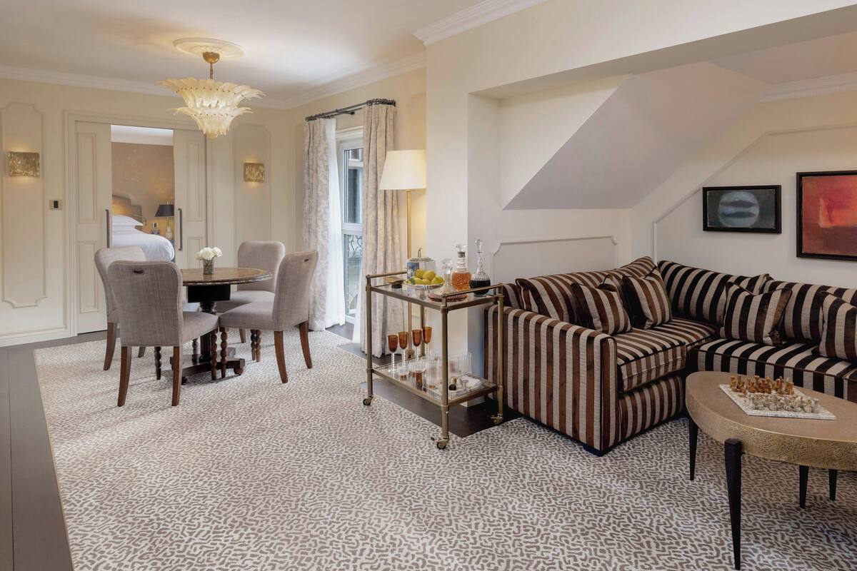 A living room with a striped sofa, a dining table with chairs, a chandelier, and a view of the bedroom.