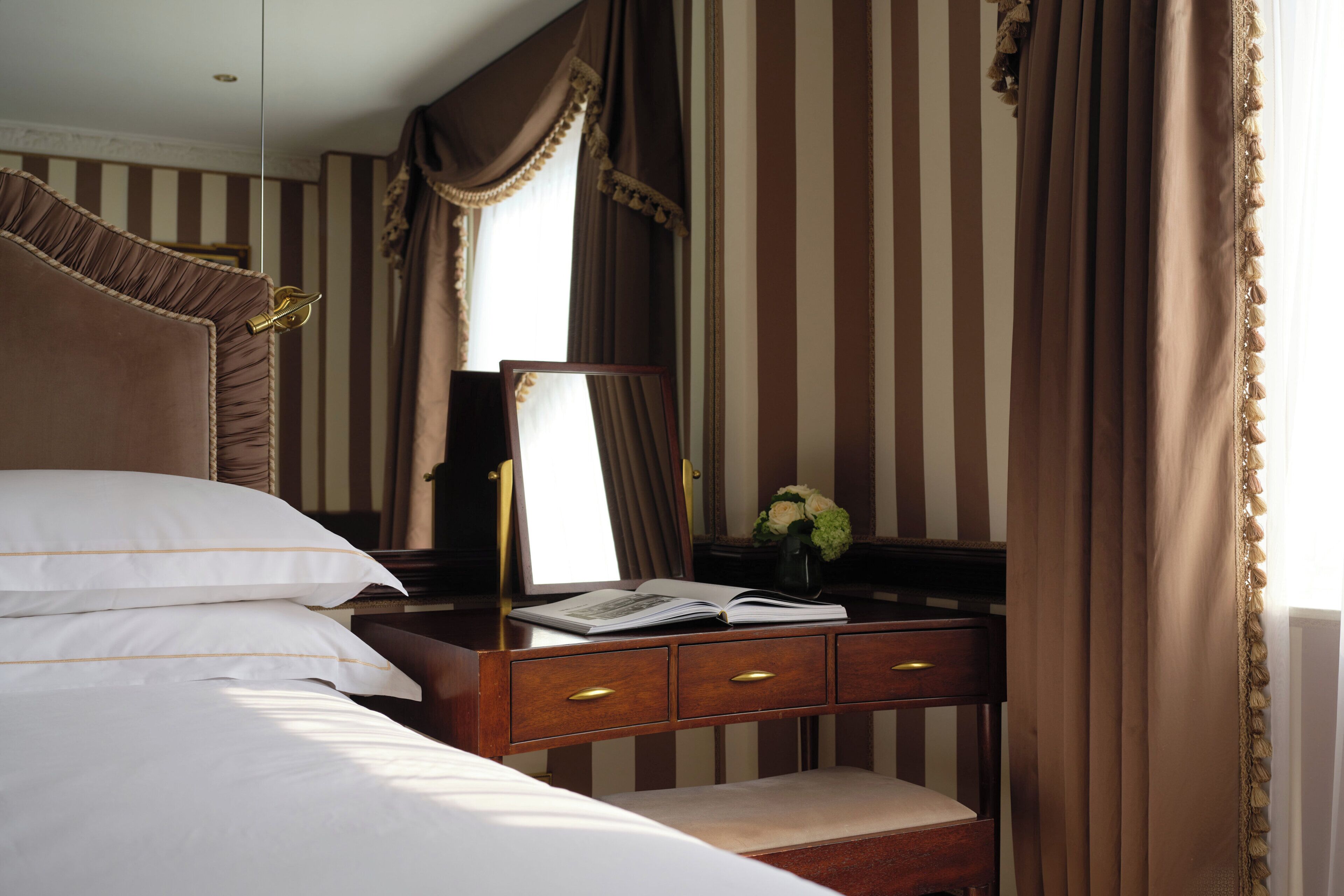 A hotel room with a bed, a bedside table with a mirror, a book, and a vase with flowers.
