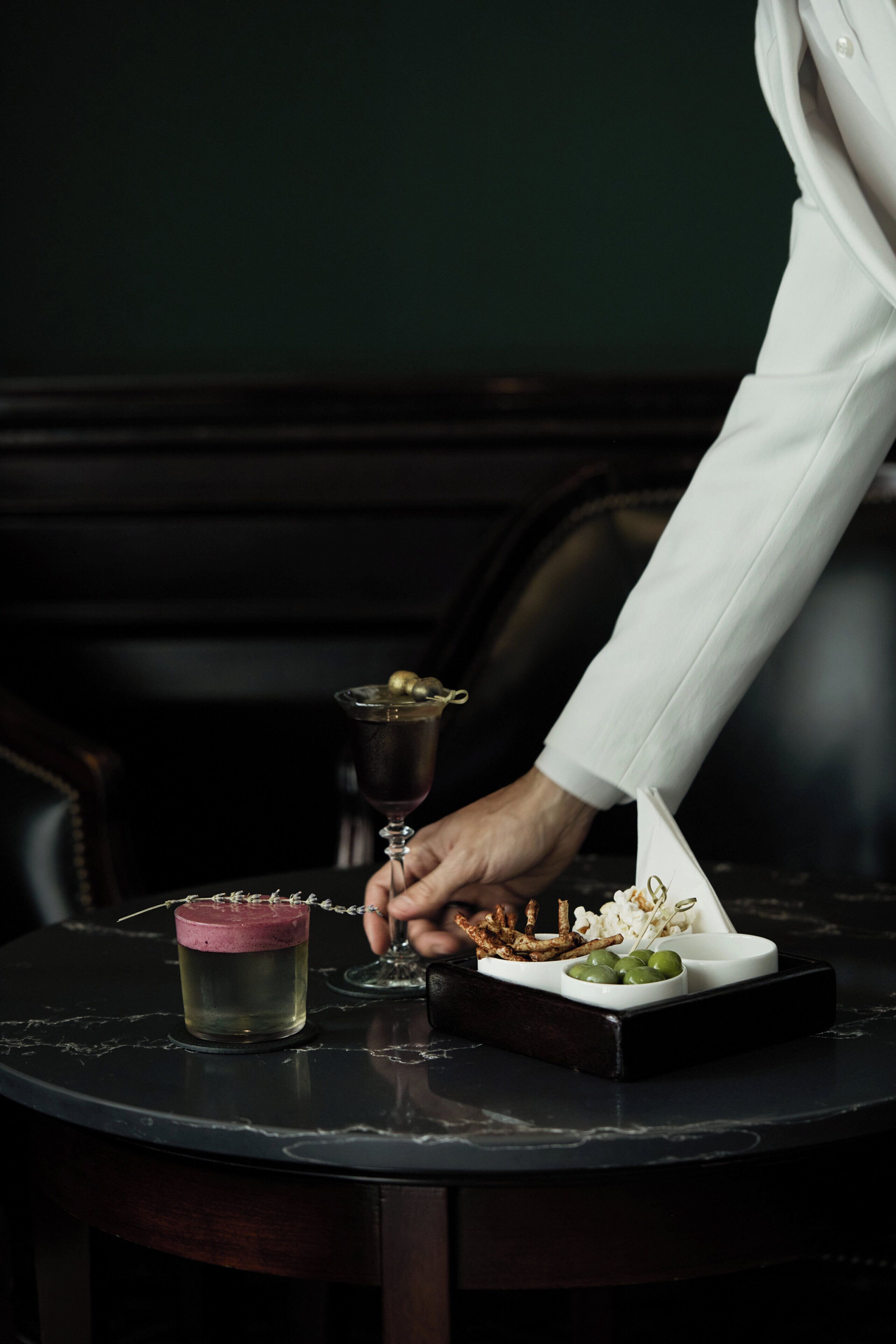 A person holding a glass with a drink, a small bowl with olives, and a dish with food on a table.