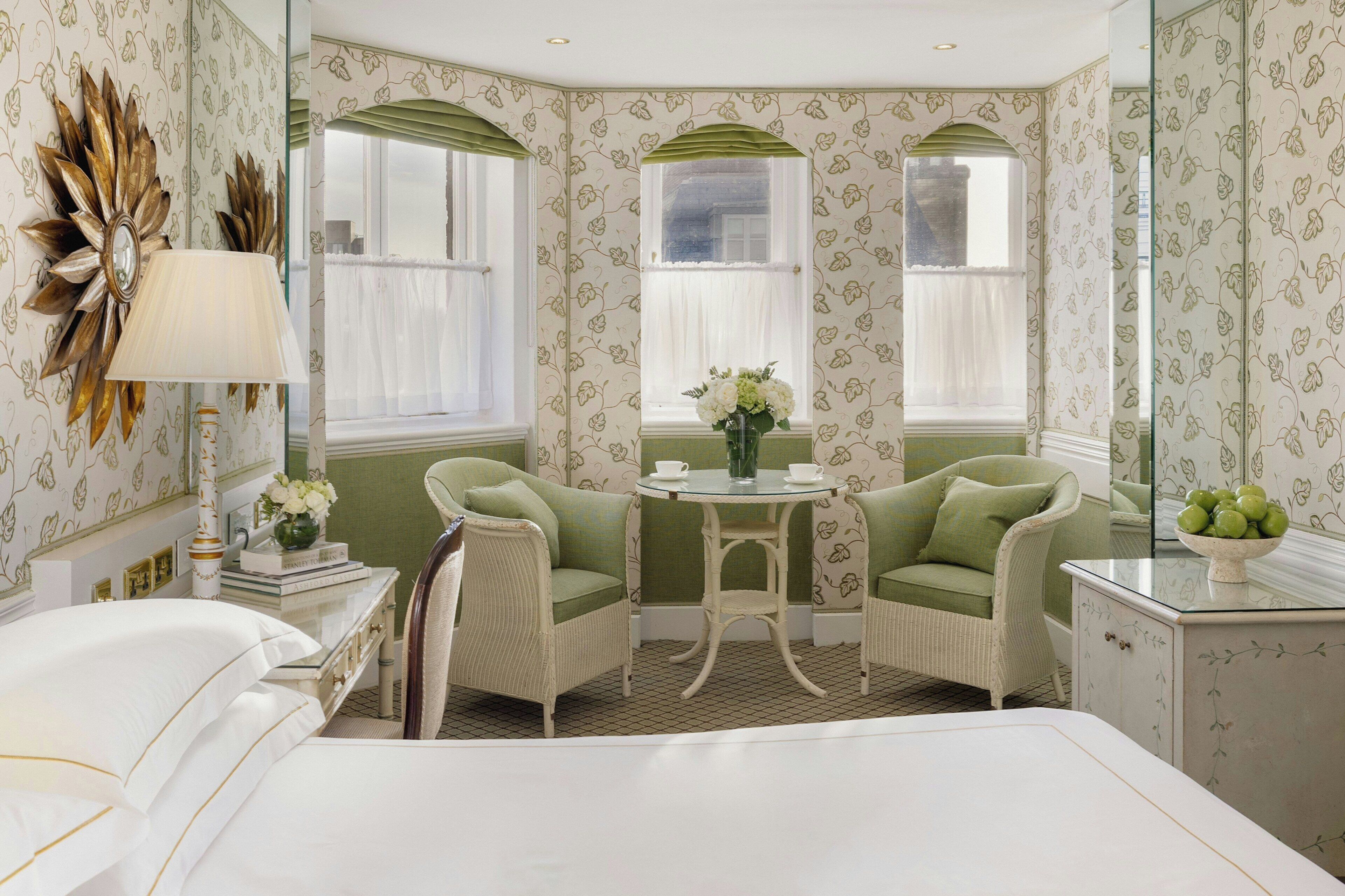 Ein Hotelzimmer mit einem Bett, zwei grünen Sessel, einem kleinen runden Tisch mit Blumenvase und einem Spiegel mit Zierrahmen.
