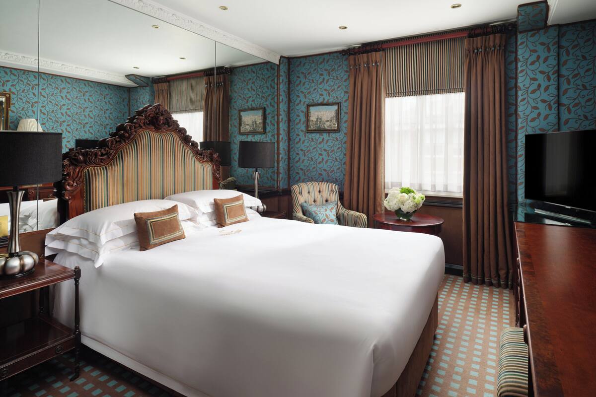 A large bed with multiple pillows, a wooden nightstand, a chair, a small table with a vase of flowers, a television, and a patterned carpet.