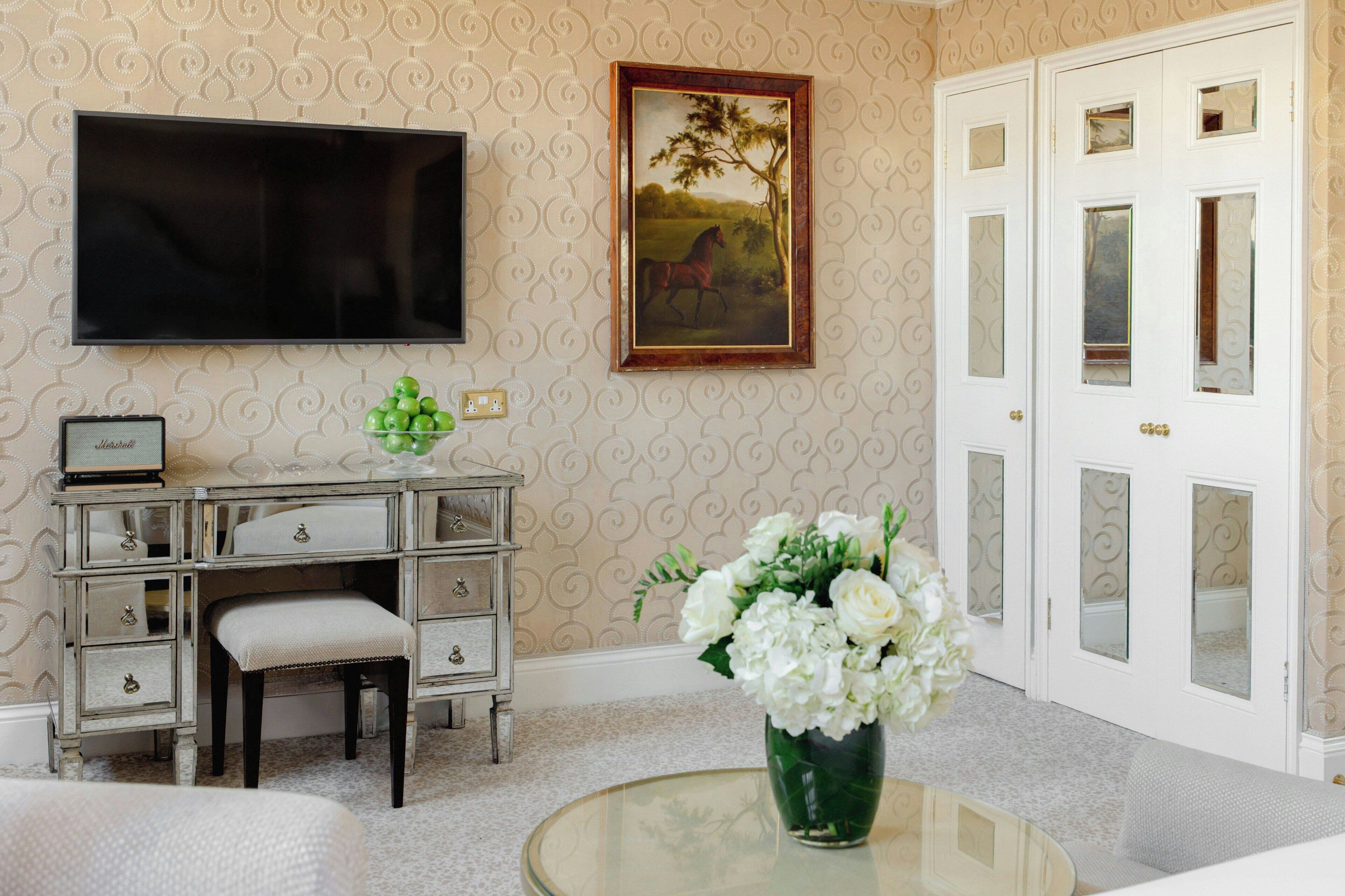 Ein Zimmer mit einem wandmontierten Flachbildfernseher, einem Schreibtisch mit Schubladen, einem Stuhl, einer Vase mit Blumen und einer Tür mit Glasfenstern.