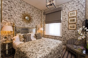 A bedroom with floral wallpaper, a large bed with multiple pillows, a bedside table with a lamp, a window with curtains, and a chair with a vase of flowers.