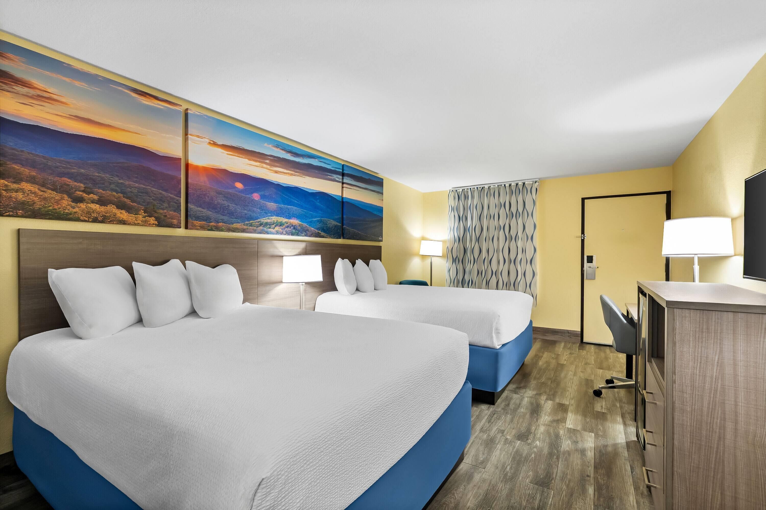 A hotel room with two beds, a desk, a lamp, and a large landscape painting on the wall.
