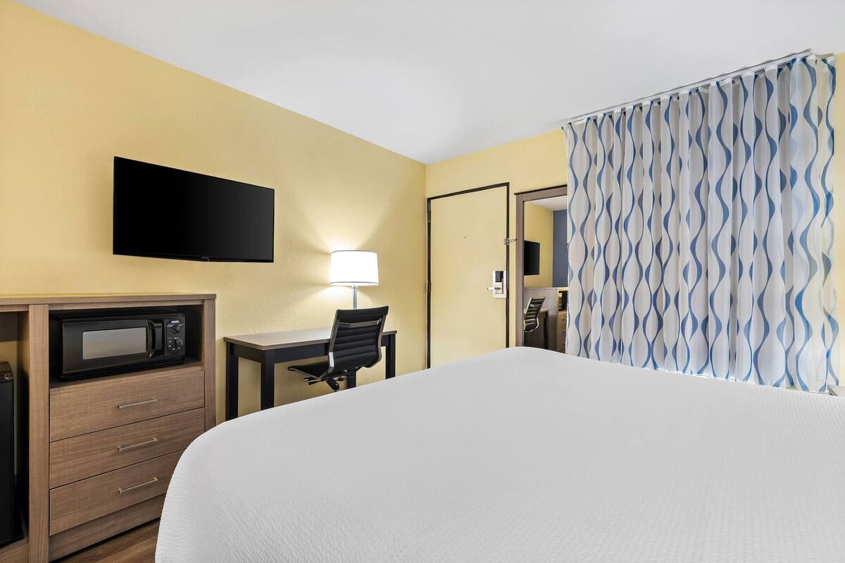 A hotel room with a bed, a desk with a lamp, a TV mounted on the wall, a microwave, and a wardrobe.