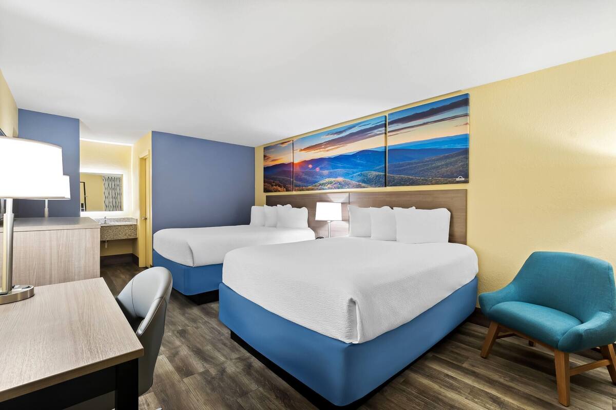 A hotel room with two beds, a desk, a chair, and a large landscape painting on the wall.