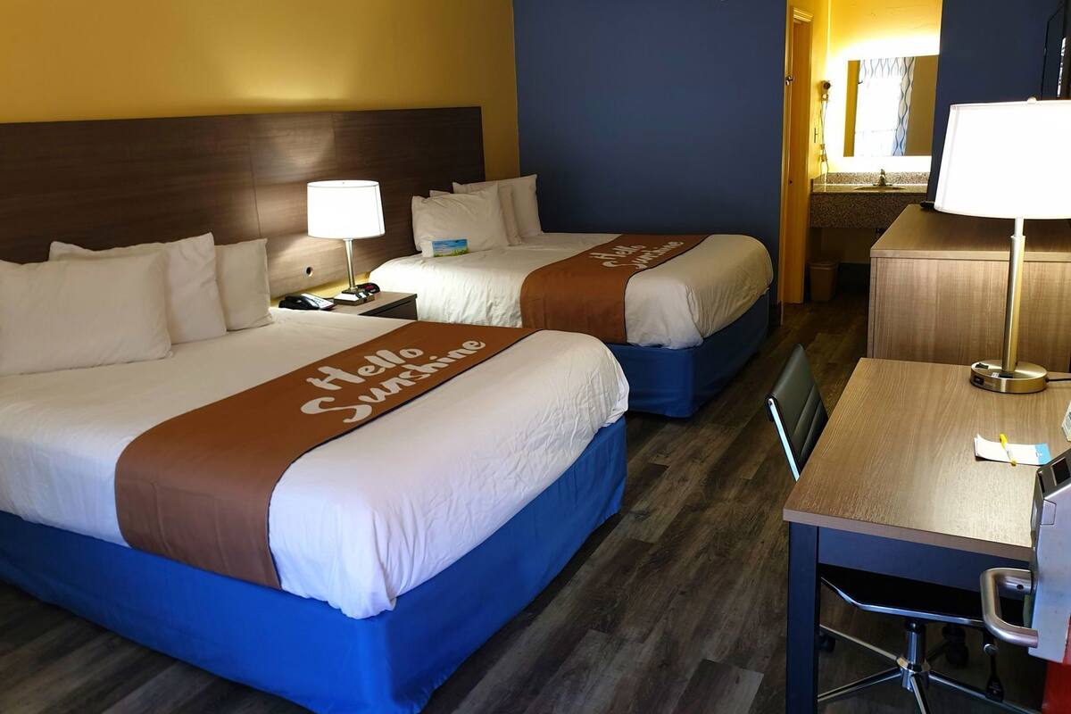 A hotel room with two beds, a desk, a chair, a lamp, and a bathroom visible in the background.