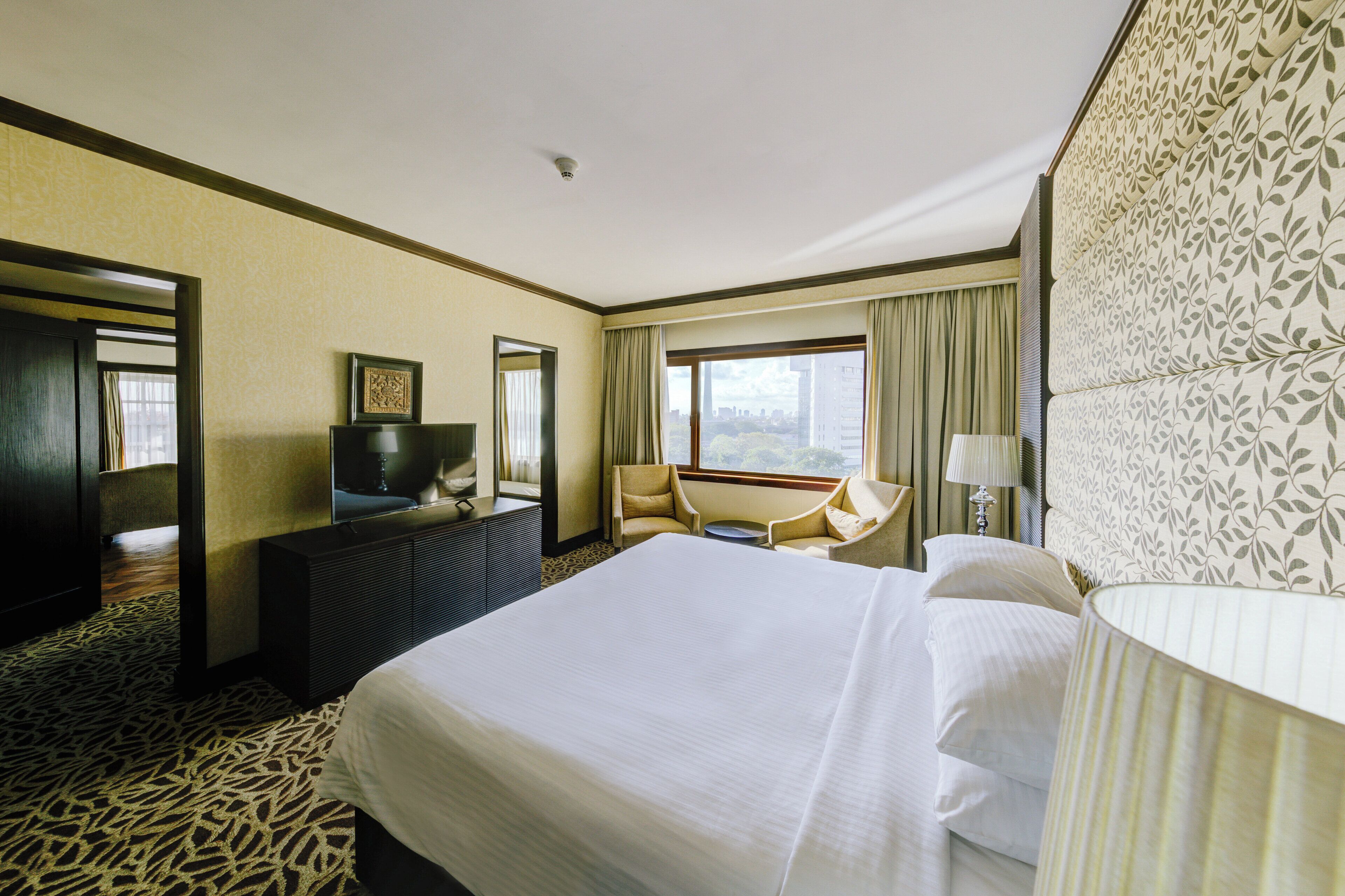 A hotel room with a large bed, a desk with a television, a chair, a window with curtains, and a patterned wall.