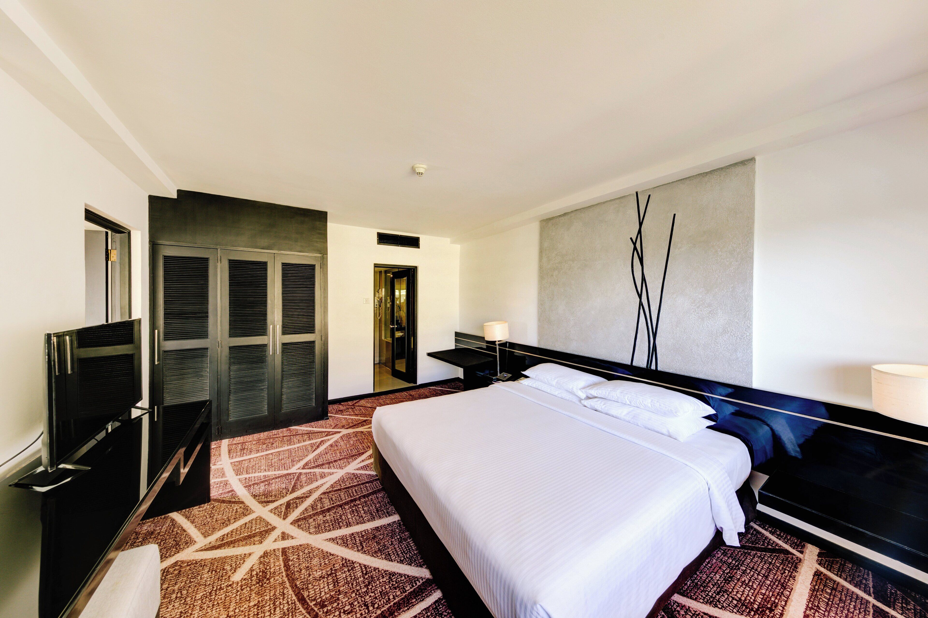 Une chambre d’hôtel dotée d’un grand lit, d’un téléviseur à écran plat, d’un bureau avec une lampe et d’une œuvre d’art contemporaine accrochée au mur.