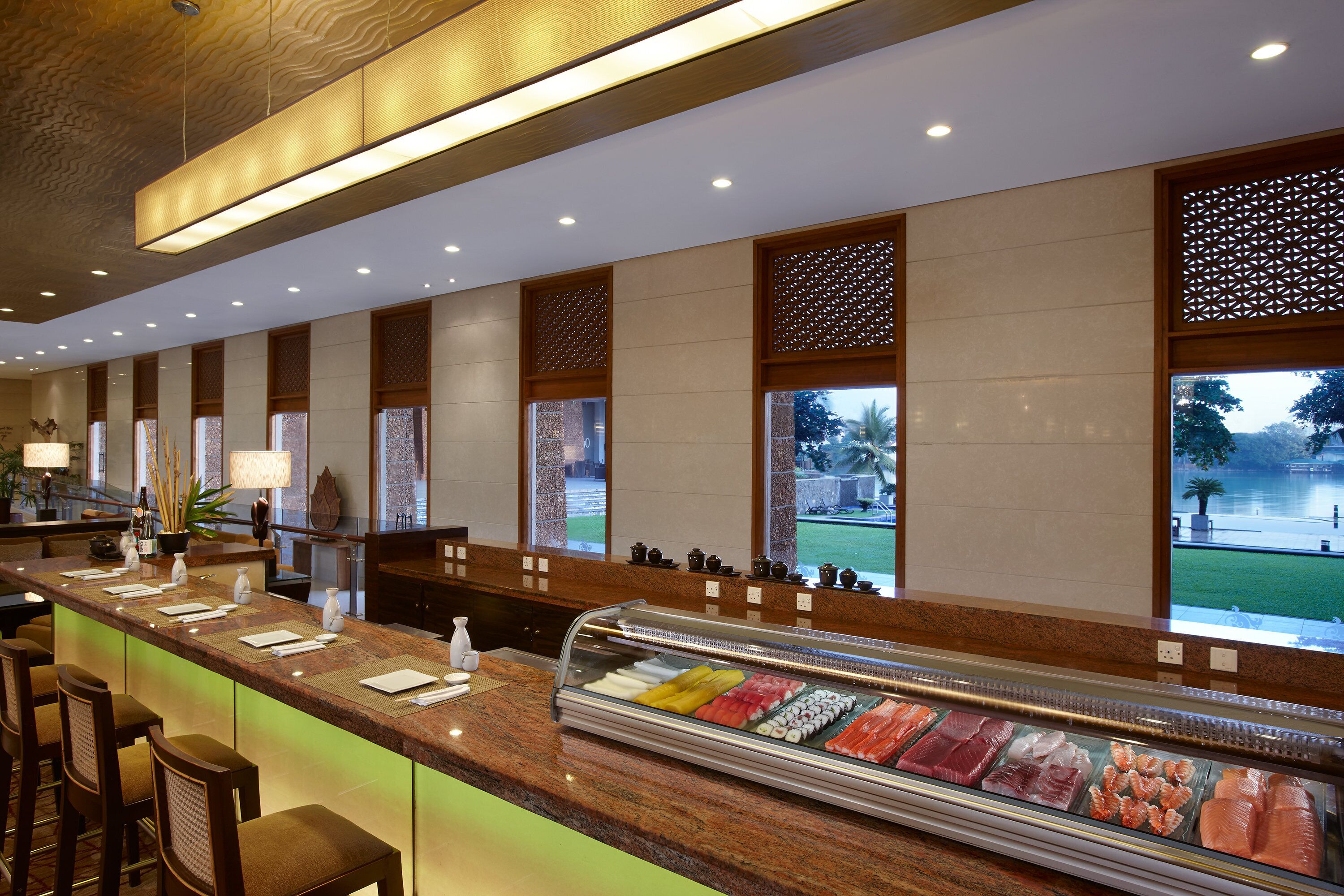 A sushi bar with a glass display case, wooden bar stools, and a view of a garden through large windows.