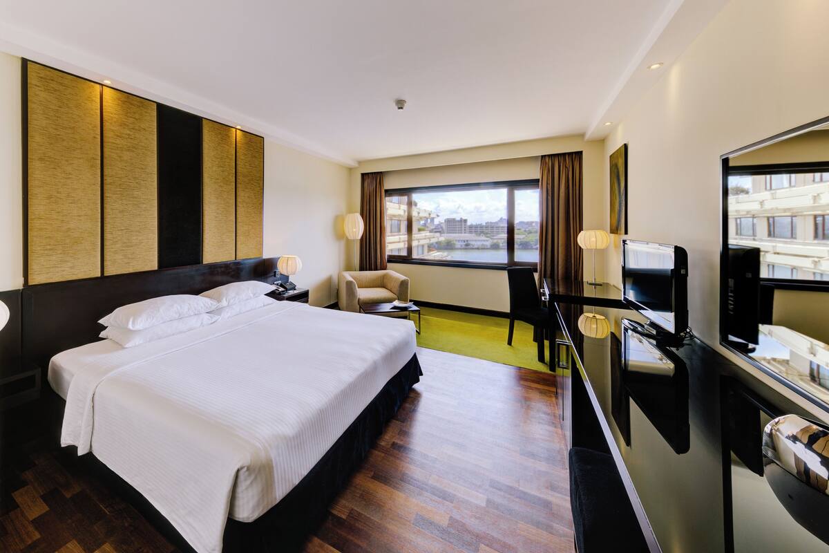 Ein Hotelzimmer mit einem großen Bett, einem Schreibtisch mit Computer, einem Stuhl, einem Fernseher und einem Fenster mit Blick auf Gebäude.