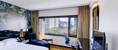 Habitación de hotel con cama, escritorio, silla, televisor y ventana grande con vista a la ciudad.