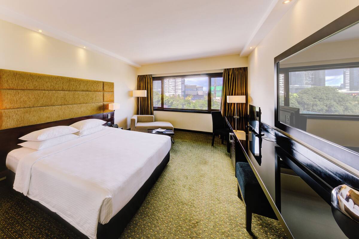 Ein Hotelzimmer mit einem großen Bett, einem Schreibtisch, einem Stuhl, einem Fernseher und einem Fenster mit Blick auf die Stadt.