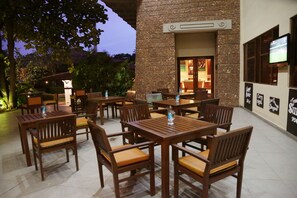 An outdoor dining area with wooden tables and chairs, a brick wall, and a television screen mounted on the wall.