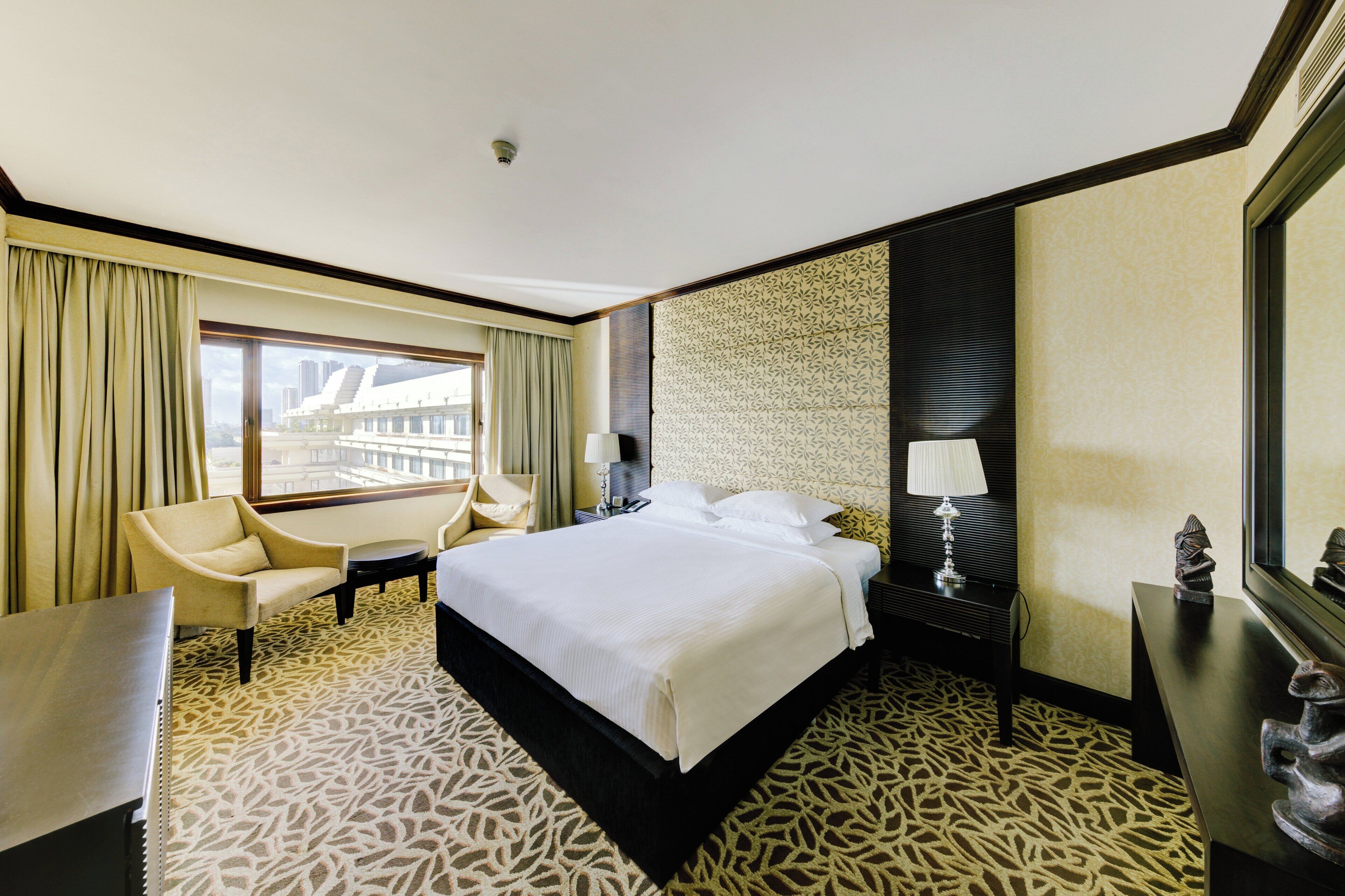 A hotel room with a large bed, a chair, a desk, and a view of a building through the window.