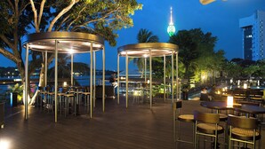 Un área de comedor al aire libre con mesas y sillas redondas, rodeada de árboles y vista al horizonte de una ciudad.