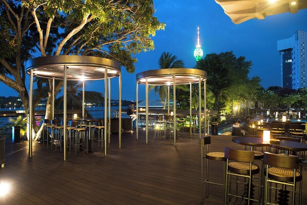 An outdoor dining area with round tables and chairs, surrounded by trees and a view of a city skyline.