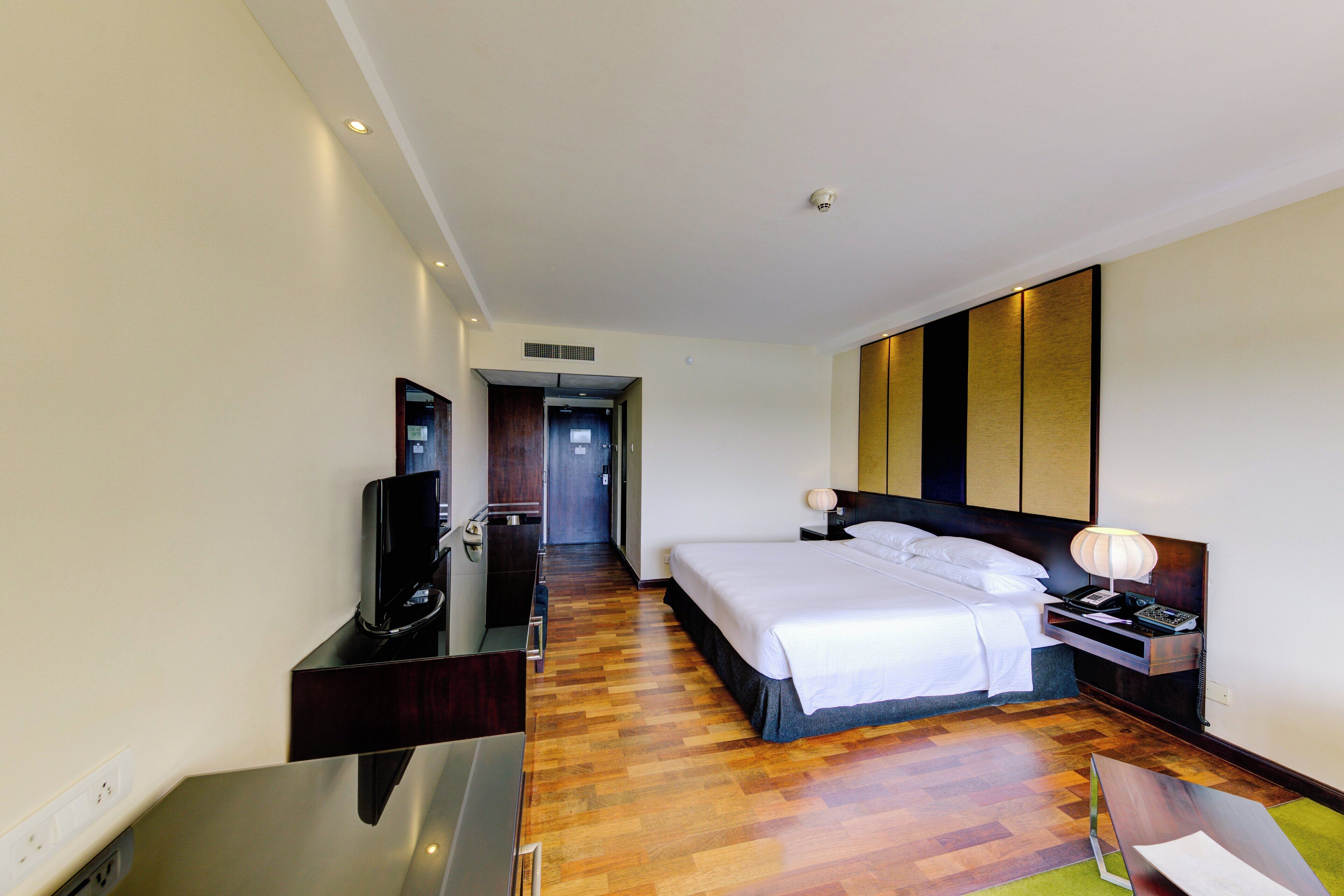 A hotel room with a bed, bedside table, lamp, TV, and a desk with a computer.