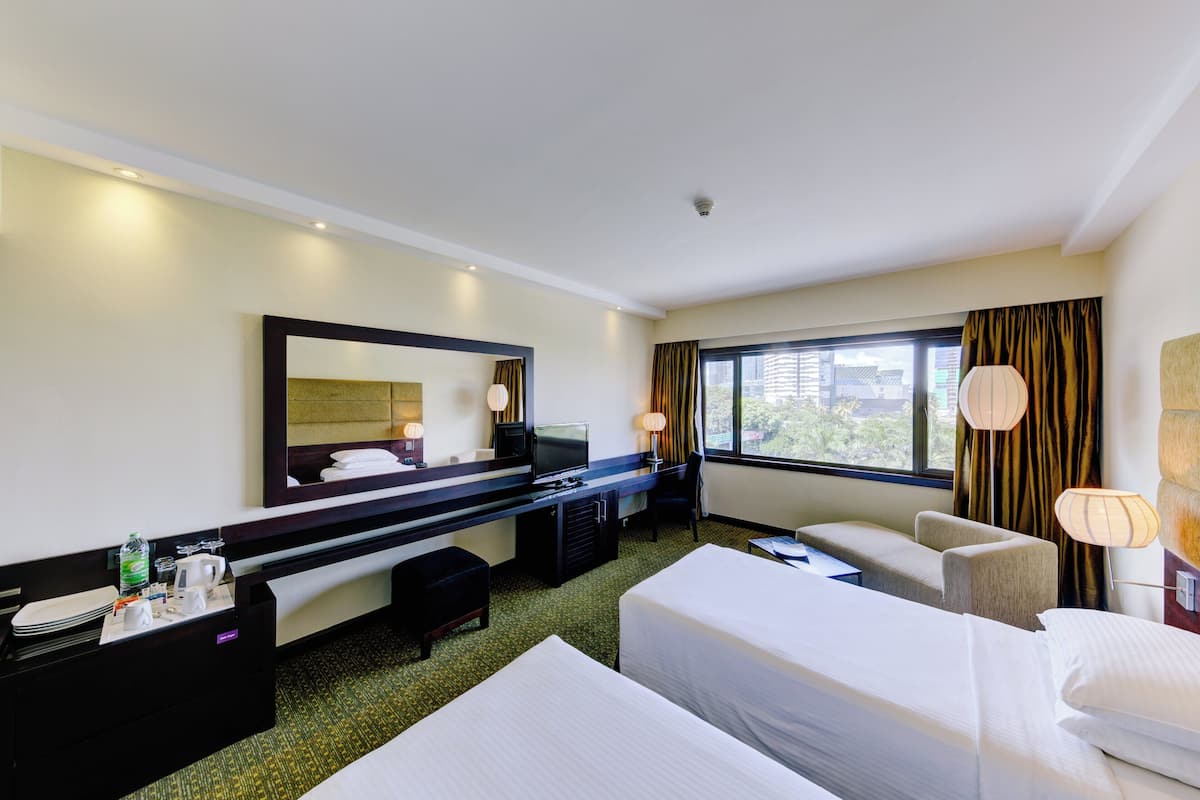 Ein Hotelzimmer mit zwei Betten, einem Schreibtisch mit Computer, einem Fernseher und einem großen Fenster mit Vorhängen.