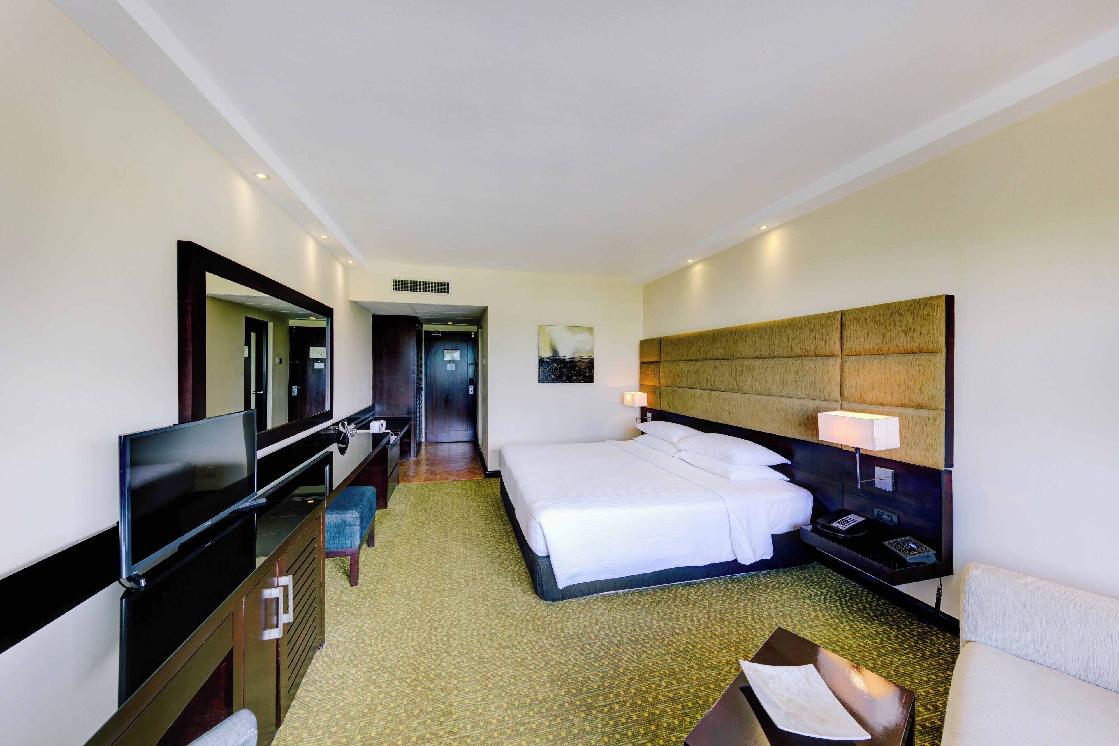 A hotel room with a large bed, a TV, a desk with a chair, and a bedside table with a lamp.