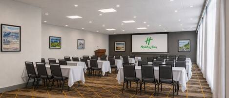 Uma sala de conferências com mesas, cadeiras, um pódio e uma tela de apresentação com o texto 'Holiday Inn'.