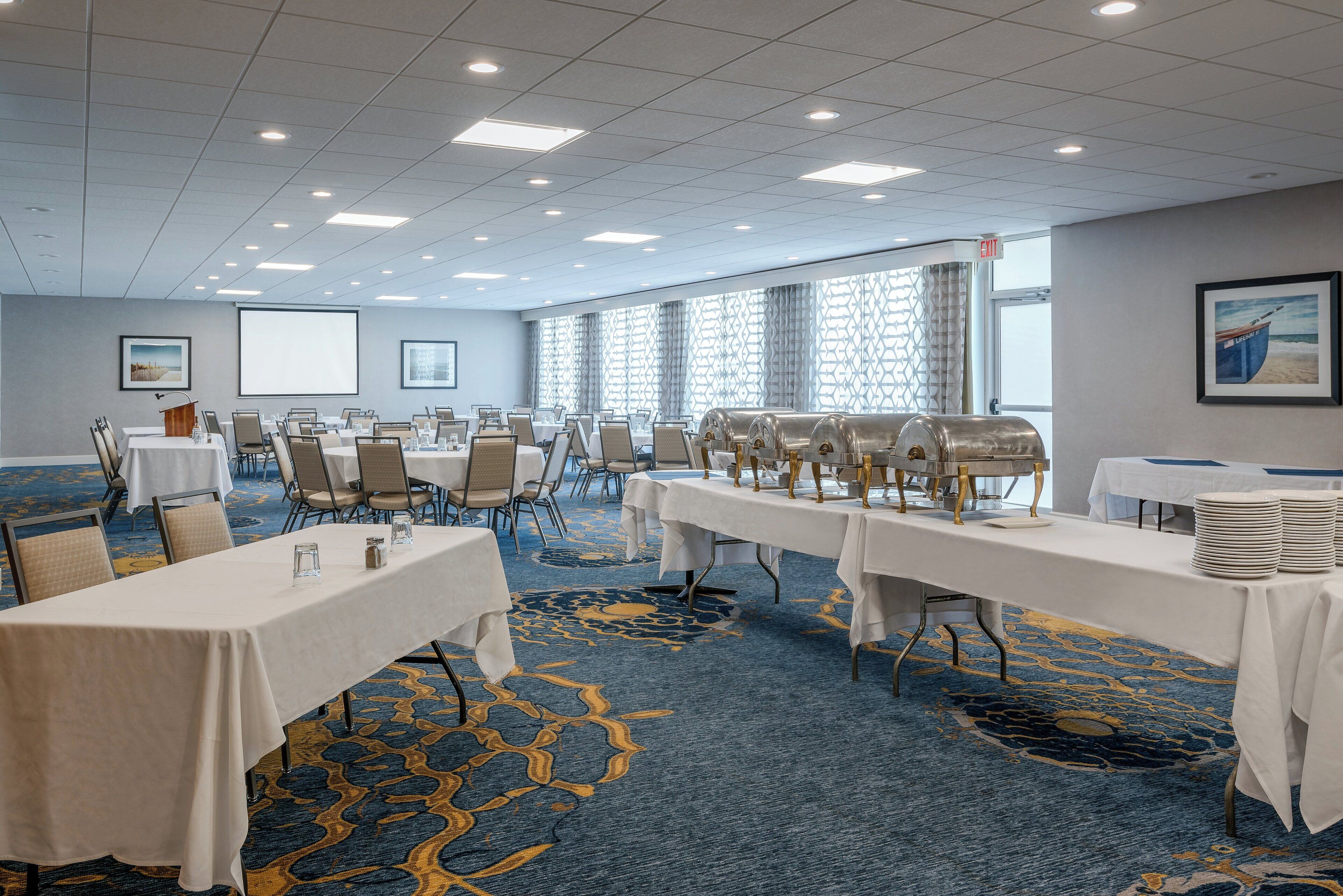 A conference room with multiple tables, chairs, a projector screen, and a buffet setup.